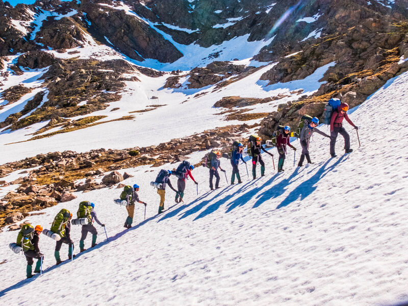 The image shows a group of people hiking up a snow-covered mountain. They are all wearing backpacks and using hiking poles. The sun is shining, and the sky is clear. The hikers are spread out along the slope, and their shadows are cast on the snow.